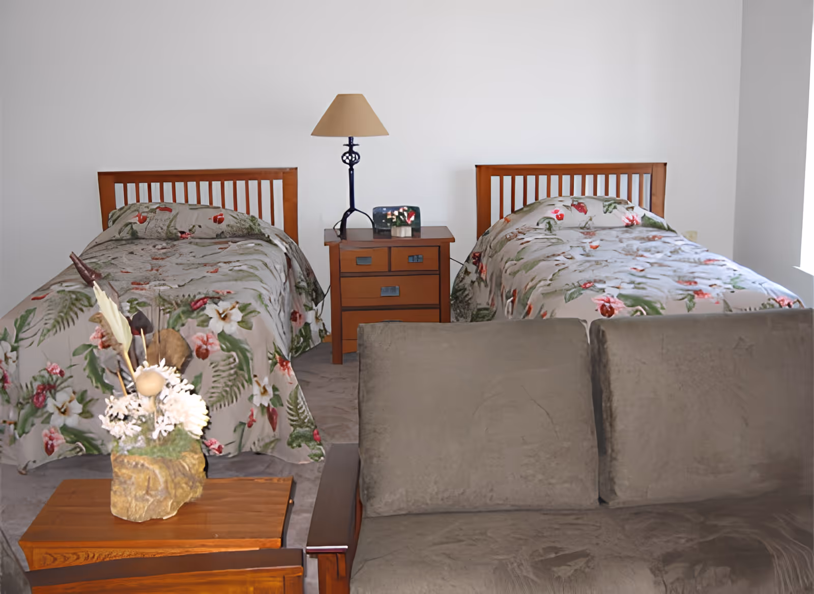 Two twin beds with floral bedspreads flanking a wooden nightstand and lamp, with a sofa and small table in the foreground.