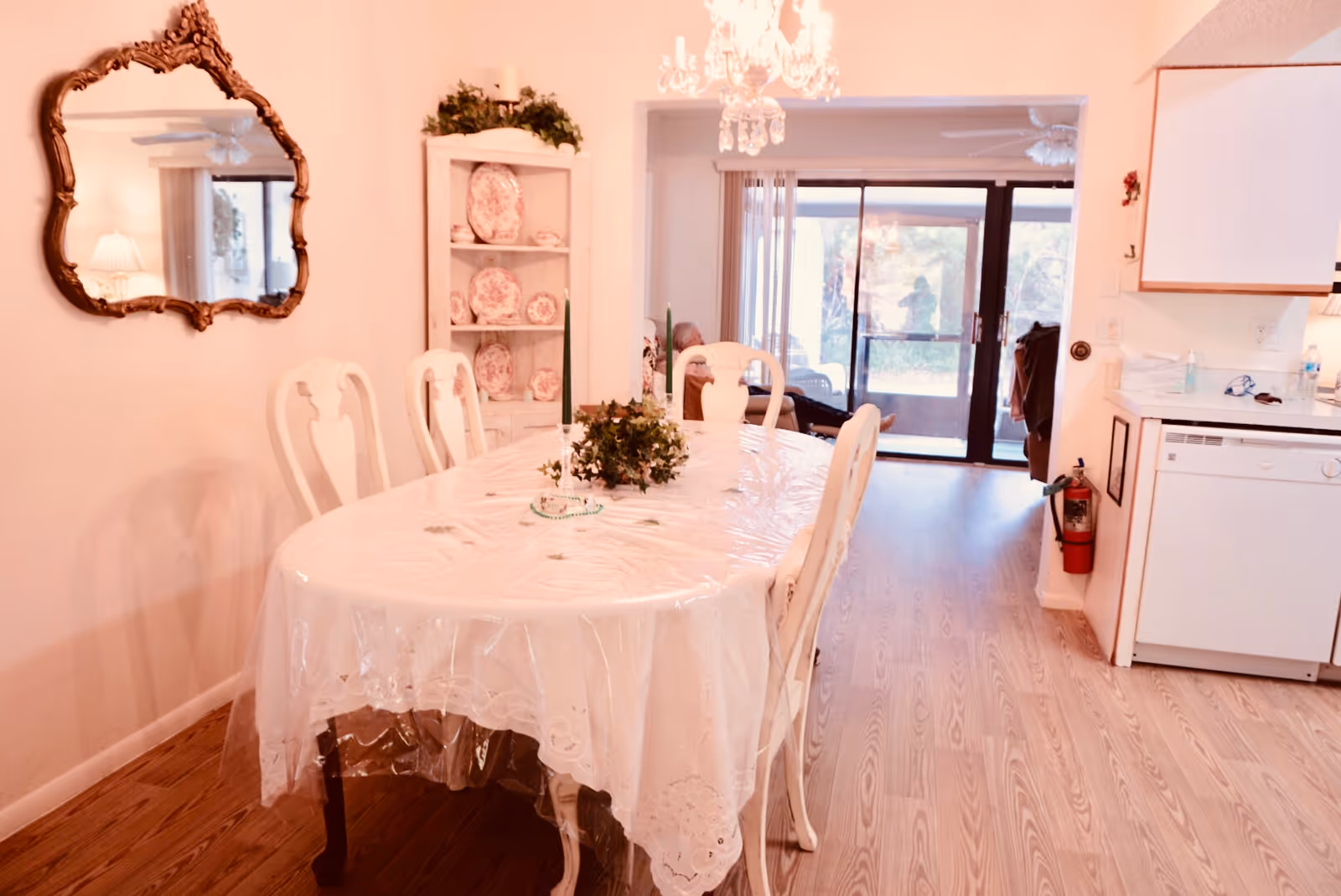 Dining room with an oval table covered in a clear plastic tablecloth, white chairs, a china hutch and chandelier, opening to sliding glass doors.