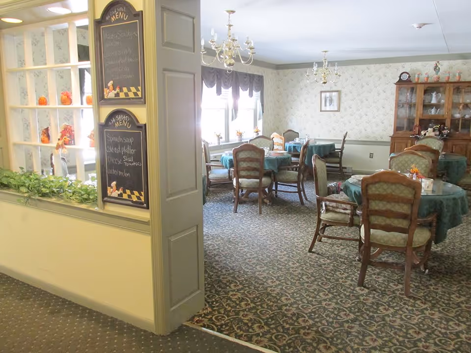 A dining room in a senior living facility with several round tables covered with green tablecloths and surrounded by wooden chairs with green upholstery. The room has patterned carpet, wallpaper with a subtle floral design, and two chandeliers hanging from the ceiling. A wooden china cabinet with dishes is visible against the far wall. Near the entrance, there are two chalkboards displaying the daily menu. A window with valances lets in natural light.