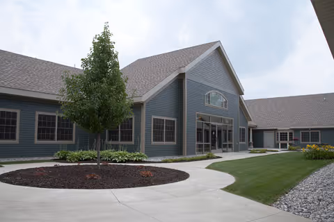 Exterior view of a single-story building with gray siding and a gabled roof. There is a concrete walkway leading to the entrance, landscaped areas with grass, a tree, and mulch. The sky is partly cloudy.