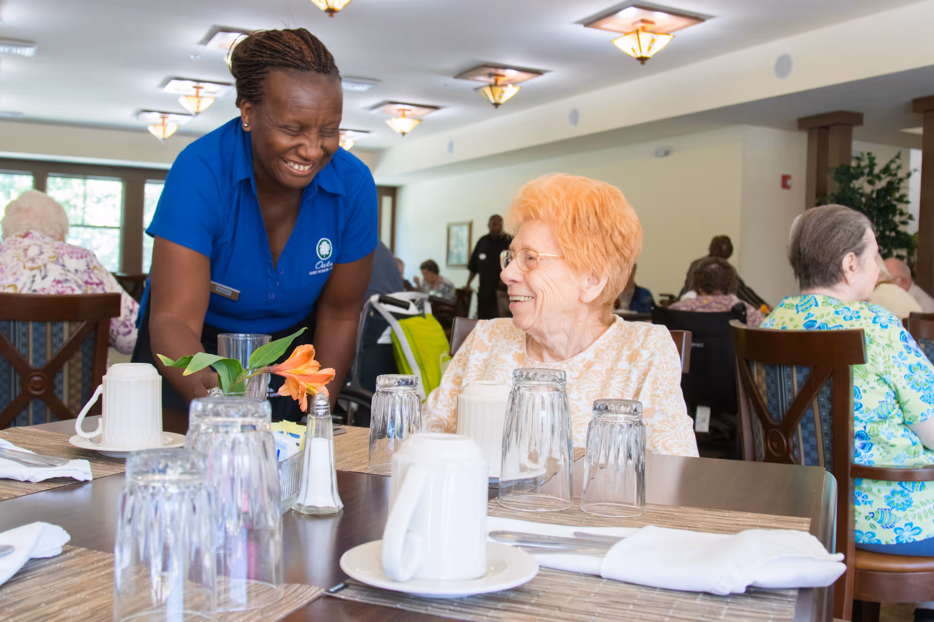 A smiling staff member in a blue shirt interacts warmly with an elderly woman with short red hair seated at a dining table set with glasses, cups, and napkins in a bright dining room with other elderly residents in the background.
