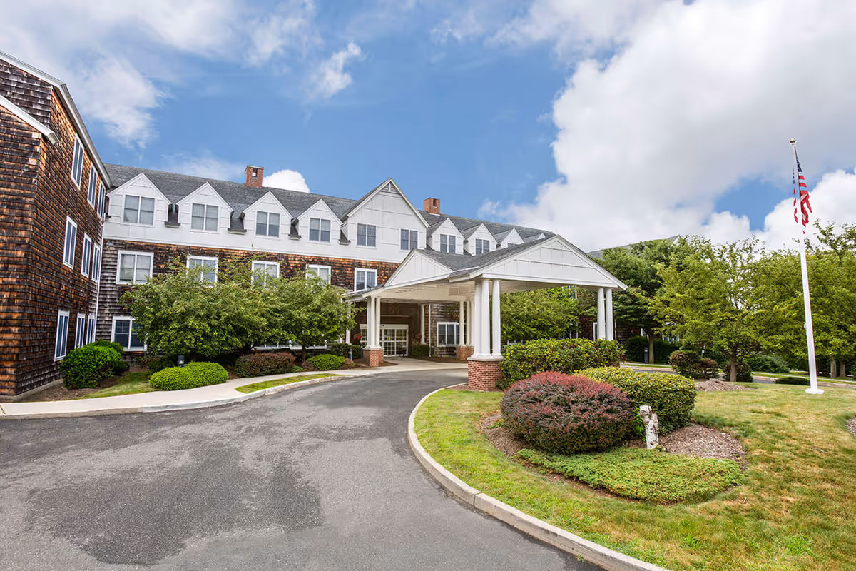 Exterior view of a senior living facility building with a covered entrance supported by white columns. The building has multiple windows and a shingled facade. There are well-maintained bushes, trees, and a lawn surrounding the driveway. An American flag on a flagpole is visible on the right side under a partly cloudy sky.