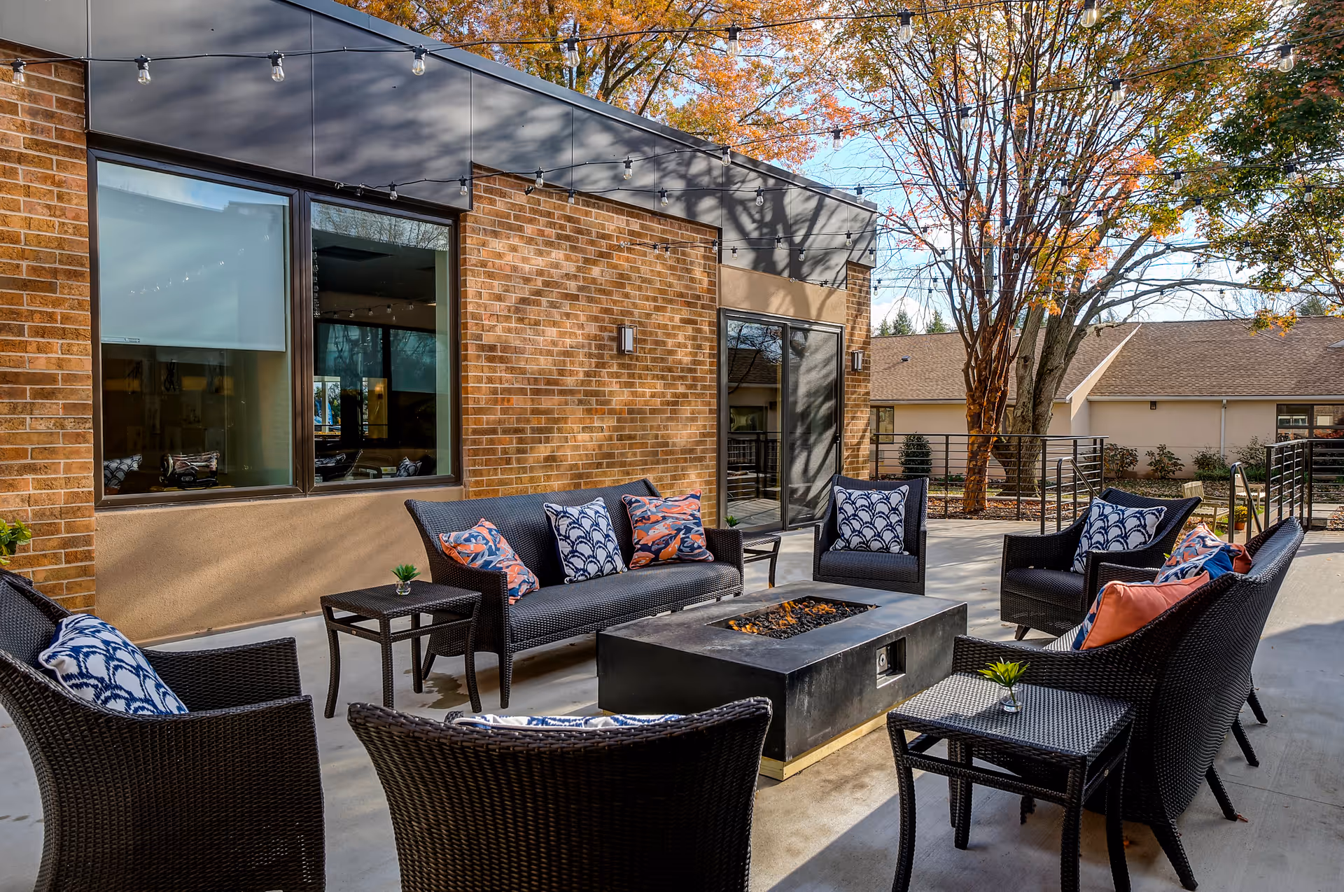 Outdoor patio area with black wicker furniture including chairs and sofas arranged around a rectangular fire pit. The seating has colorful patterned cushions. The patio is adjacent to a brick building with large windows and a glass door. Trees with autumn foliage are visible in the background.