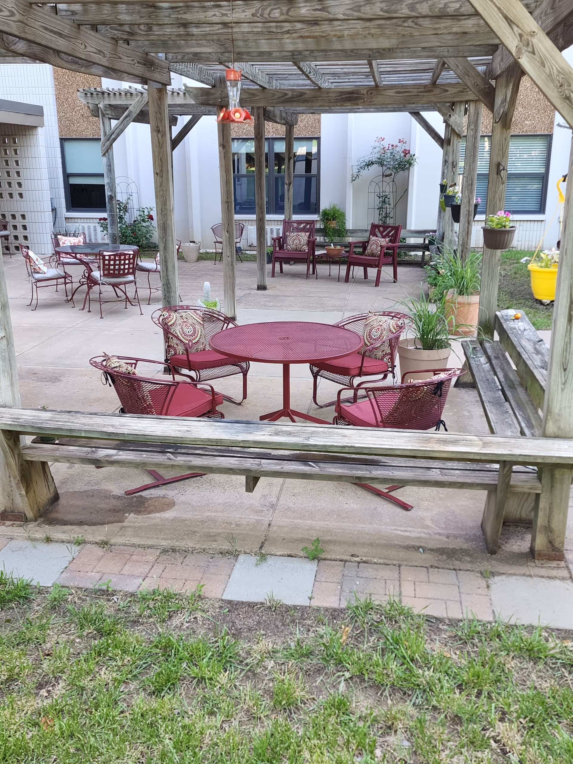 Outdoor patio area at Arkansas City Presbyterian Manor with a wooden pergola overhead. There are several red metal tables and chairs with patterned cushions arranged on a concrete surface. Potted plants and hanging flower pots decorate the space. The building exterior with windows is visible in the background.