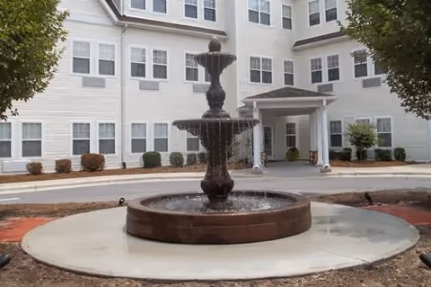 Three-tiered fountain in front of a multi-story white senior living building entrance with many windows.