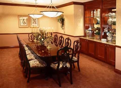 A long wooden dining table with upholstered chairs, a chandelier, and a display cabinet of china.