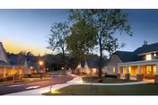 Exterior view of a senior living facility at dusk with a paved walkway, street lamps, trees, and multiple single-story buildings with lit windows and porches.