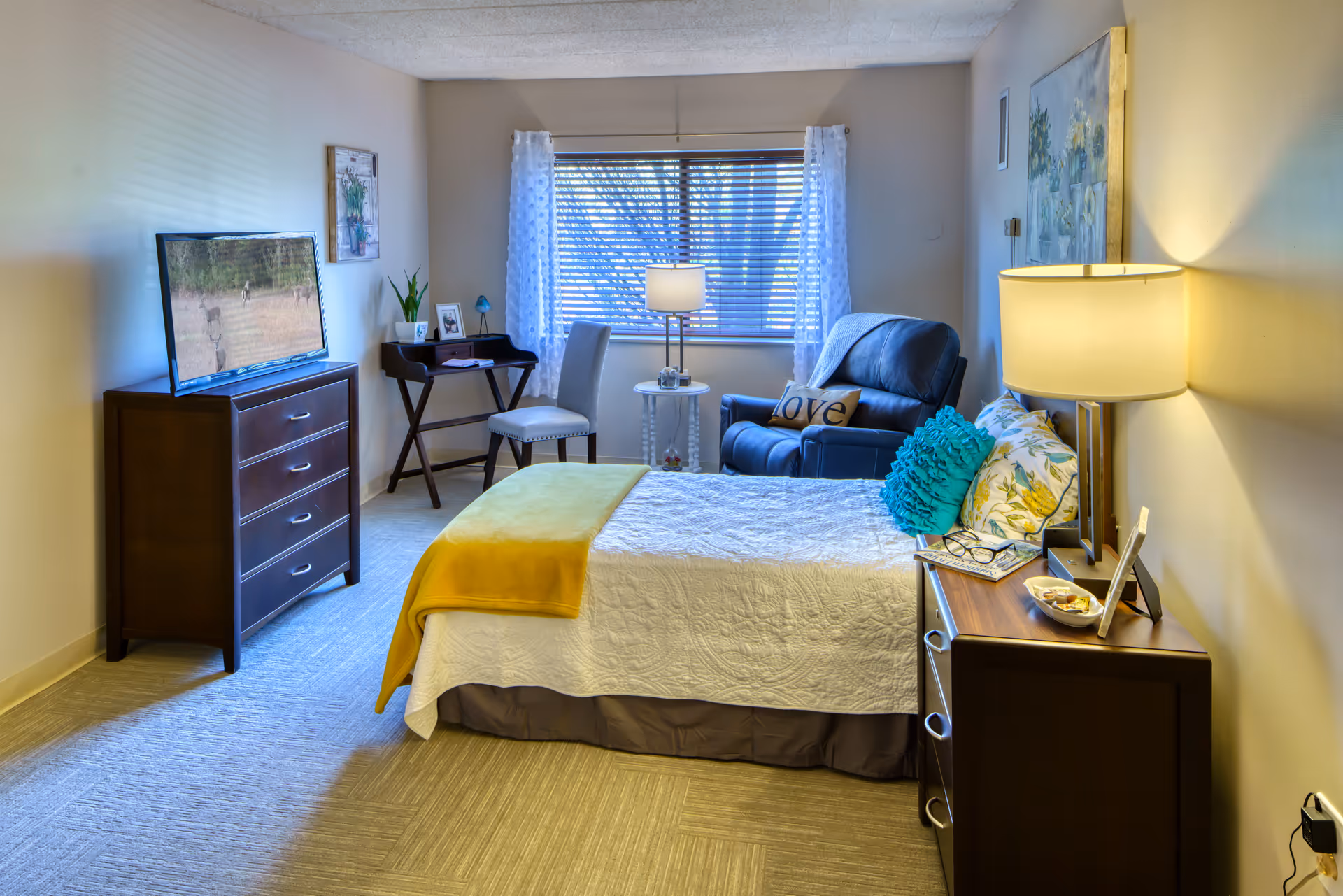Furnished senior living bedroom with a bed, dresser and TV, armchair by the window and bedside tables with lamps.
