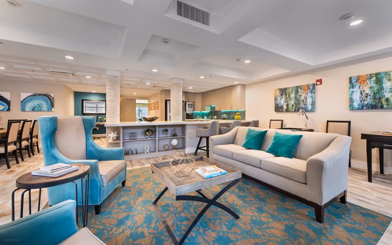 Bright communal living room with a beige sofa, teal accent chairs, a coffee table on a patterned rug, and a bar/kitchenette in the background.