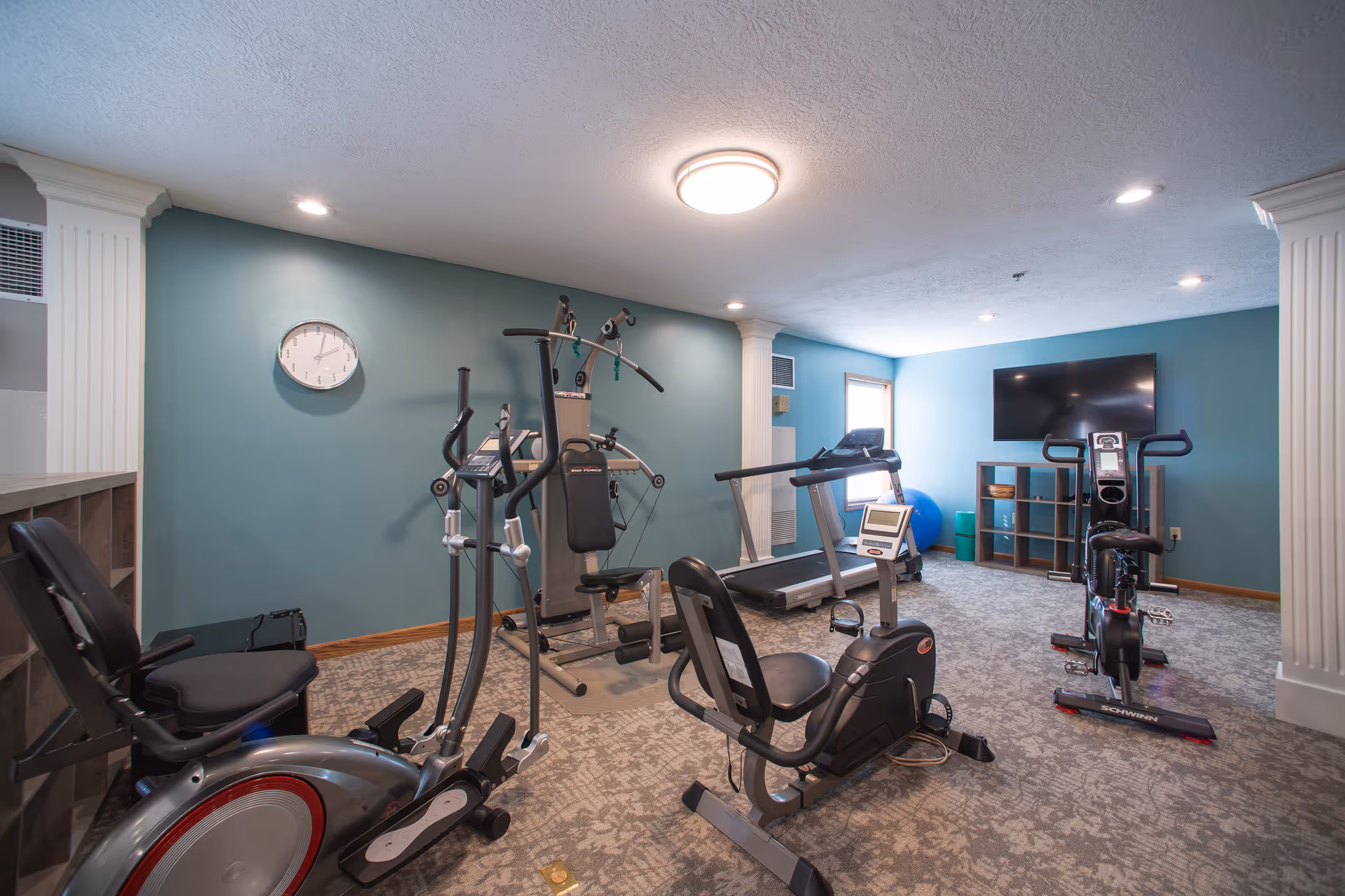 A small fitness room with various exercise equipment including a recumbent bike, elliptical machine, multi-gym, treadmill, and upright bike. The room has teal walls, a wall clock, a mounted flat-screen TV, and a window letting in natural light. The floor is carpeted and the ceiling has recessed lighting and a central light fixture.