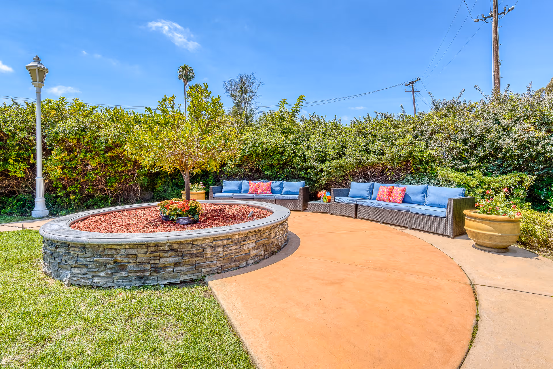Outdoor courtyard with a circular raised stone planter, wicker sofas with blue cushions and colorful pillows, potted plants and a lamppost under a clear sky.