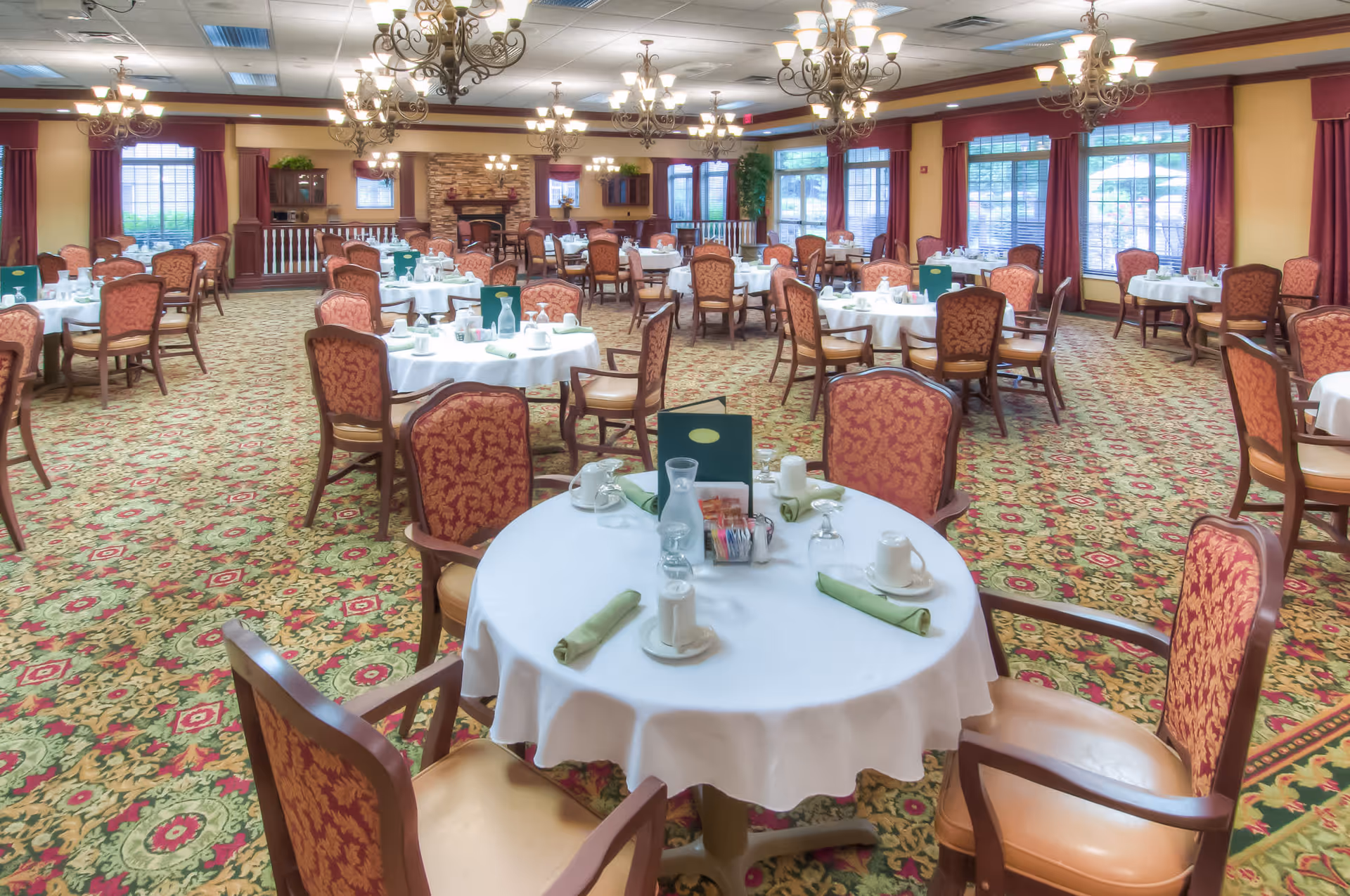 A spacious dining room with multiple round tables covered with white tablecloths, each set with cups, glasses, green napkins, and menus. The room features patterned carpet, red upholstered chairs, large windows with red curtains, and several ornate chandeliers hanging from the ceiling. There is a stone fireplace and wooden cabinetry in the background.