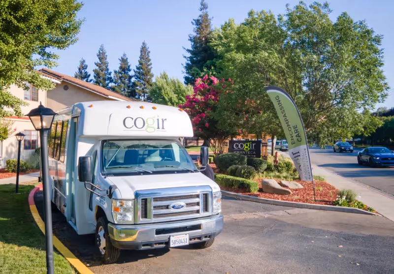 A white shuttle bus with the logo 'cogir' parked in front of the Cogir of Manteca senior living facility. The bus is parked on a curved driveway next to a landscaped area with trees, bushes, and a flag banner that reads 'ENCOMPASSING CARE'. The building and a street with cars are visible in the background under a clear blue sky.