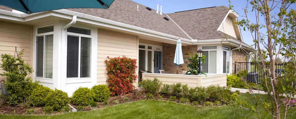 Exterior view of a single-story senior living unit with a covered patio, umbrella, and manicured landscaping.