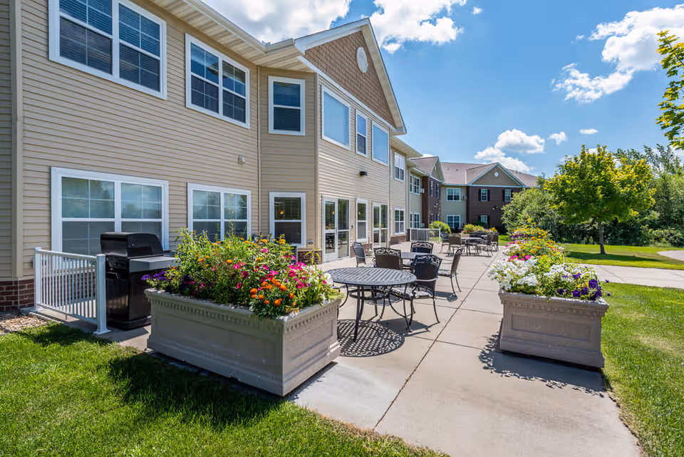 Outdoor patio area at Edgewood Sartell with metal tables and chairs arranged on a concrete walkway. Large planters filled with colorful flowers line the patio. The building exterior is beige with multiple windows, and there is a grill near the building. Green grass and trees surround the patio under a partly cloudy blue sky.