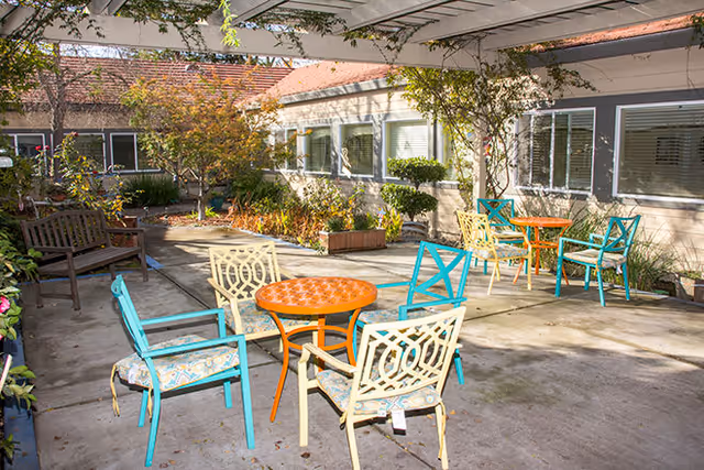 Outdoor patio area with colorful metal chairs and round tables under a pergola. The patio is surrounded by plants, shrubs, and a building with multiple windows.