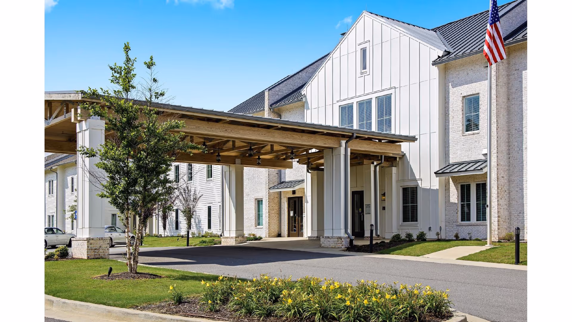 Covered porte-cochere and landscaped driveway at the front entrance of a white senior living building with an American flag.
