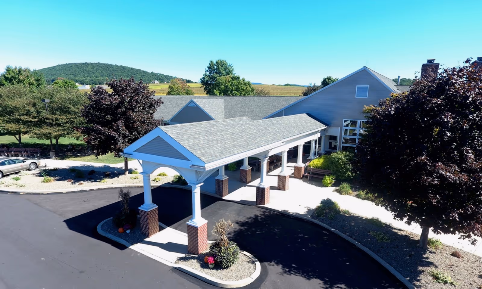Exterior view of The Meadows, A Park Home Personal Care Community, showing the entrance with a covered driveway supported by white columns with brick bases, surrounded by trees and landscaped areas, with hills and fields in the background under a clear blue sky.
