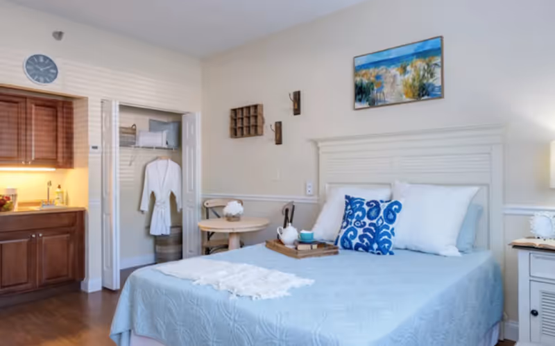 A cozy bedroom with a light blue bedspread, white and blue pillows, and a wooden headboard. There is a small round table with two chairs next to the bed, a tray with a teapot and cups on the bed, and a closet with a white robe hanging inside. The room has wooden flooring, a kitchenette with cabinets and a sink, a wall clock, and a painting of a beach scene above the bed.