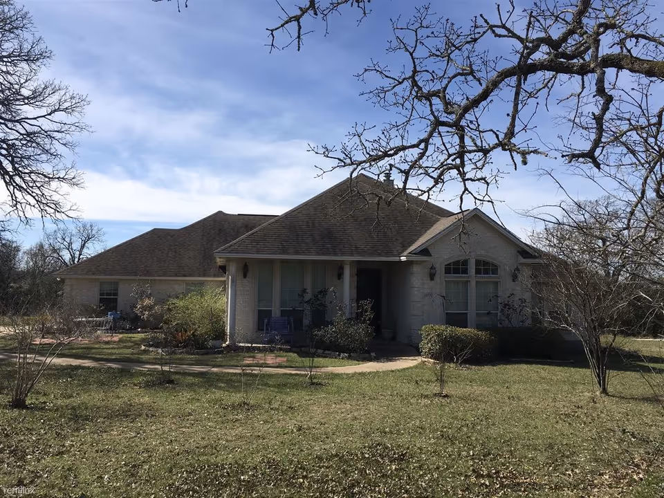 Single-story house with a sloped roof, light-colored brick exterior, and a front porch with two chairs. The house is surrounded by a grassy yard with some bushes and leafless trees under a partly cloudy blue sky.