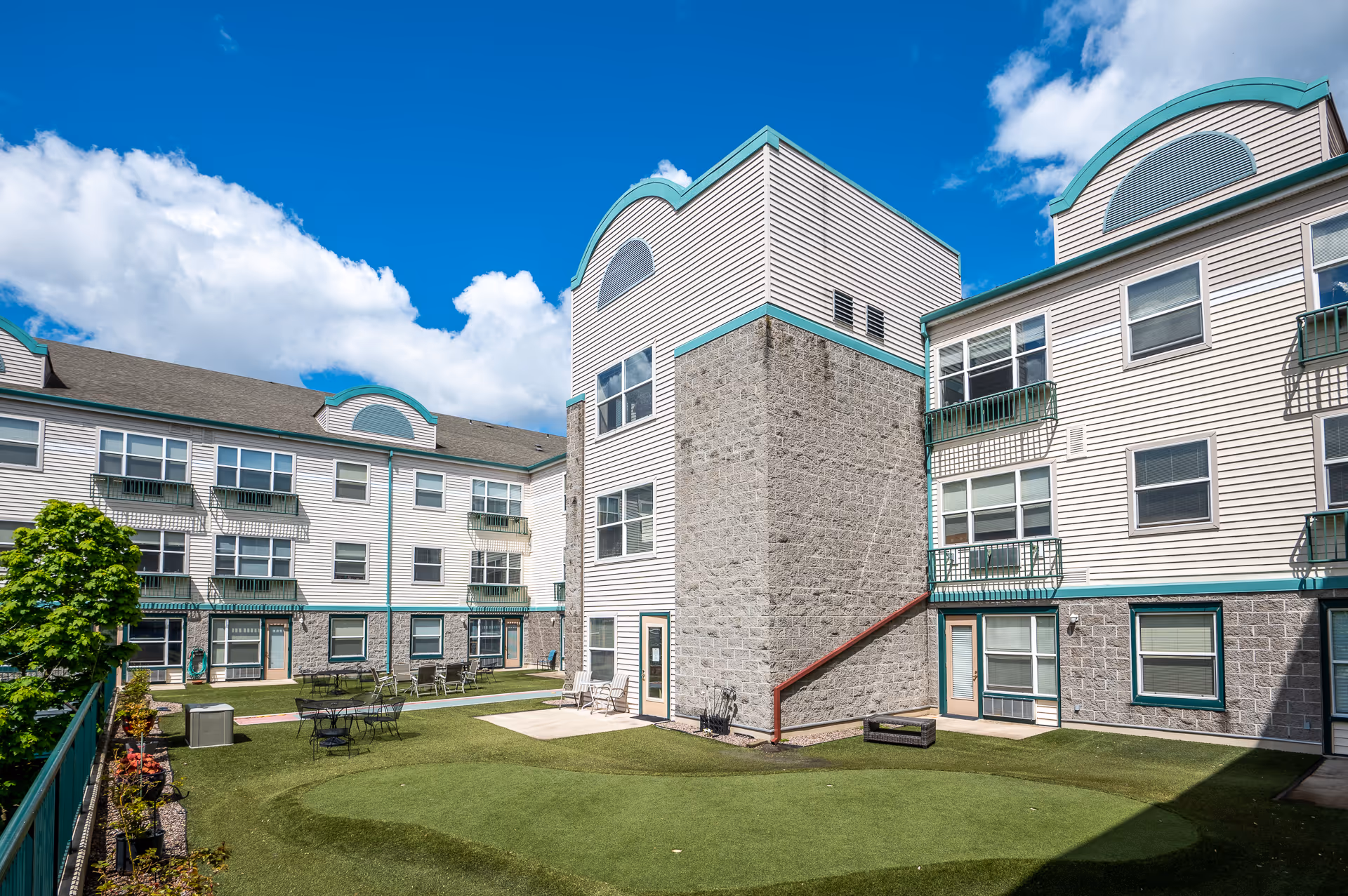 Outdoor courtyard area of Hazelwood Community Apartments featuring a putting green, patio tables and chairs, surrounded by a three-story building with white siding and green trim under a blue sky with some clouds.