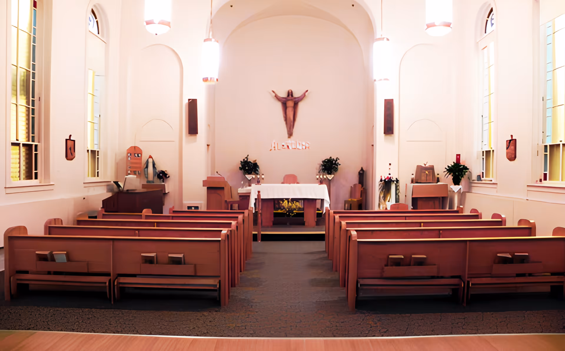 Interior view of a chapel with wooden pews arranged in rows facing an altar. The altar is covered with a white cloth and decorated with plants and candles. A crucifix is mounted on the wall behind the altar. The chapel has tall arched windows on both sides and hanging light fixtures.