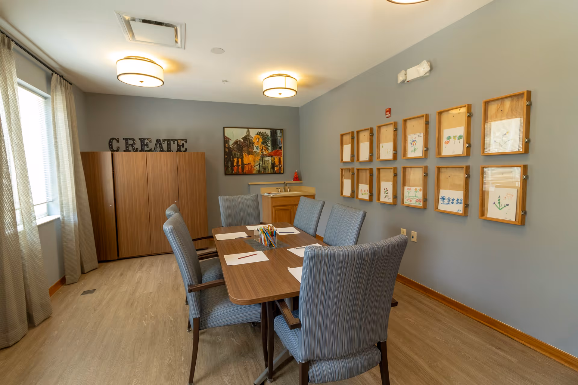 A well-lit room with a wooden table surrounded by six striped upholstered chairs. On the table are sheets of paper and colored pencils in a container. The walls are painted gray, with framed artwork and drawings displayed on one wall. A wooden cabinet with the word 'CREATE' on top is against another wall, next to a window with light curtains. Ceiling lights illuminate the room.