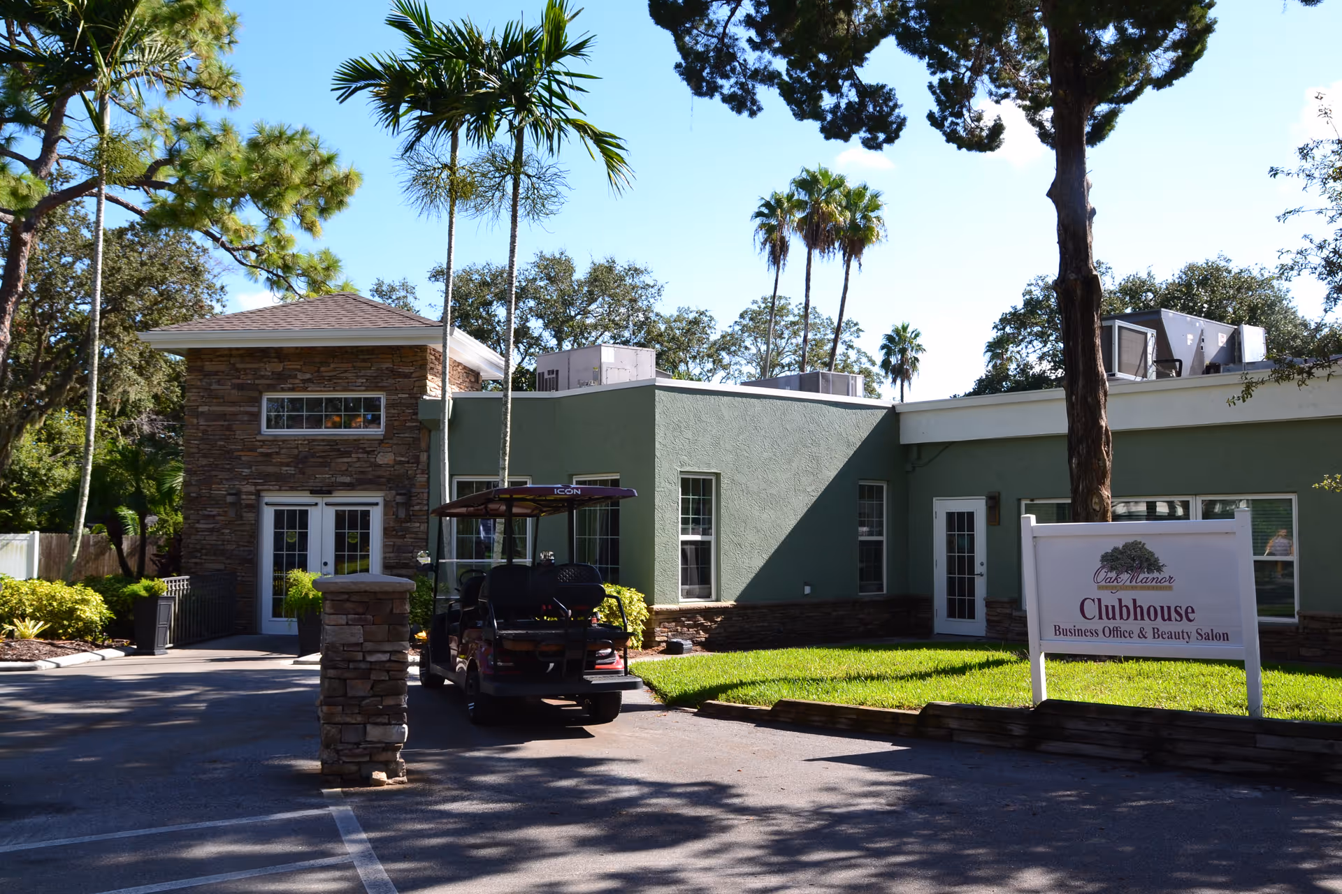 Exterior view of Oak Manor Senior Living Community's clubhouse building with green walls and stone accents. There is a golf cart parked in front, surrounded by trees and greenery under a clear blue sky. A sign in front reads 'Oak Manor Clubhouse Business Office & Beauty Salon.'