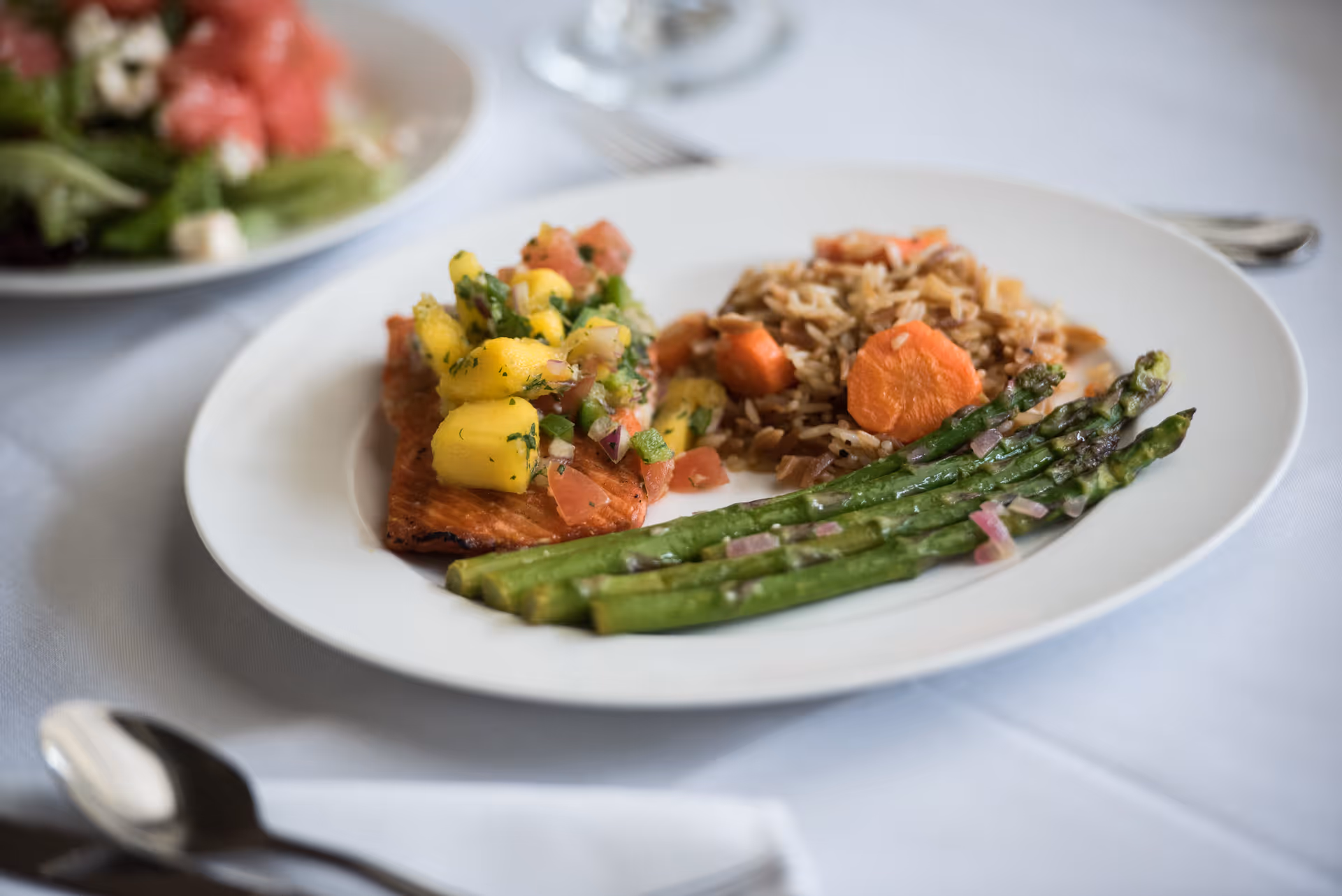 A white plate with a serving of grilled salmon topped with mango salsa, a side of cooked rice with carrots, and grilled asparagus. In the background, there is a blurred salad plate on a white tablecloth with silverware nearby.