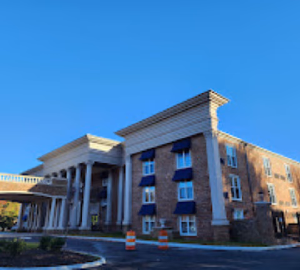 Exterior view of a multi-story brick building with white columns and blue window awnings under a clear blue sky. The building has a classical architectural style with a covered entrance and a driveway with two orange traffic cones.