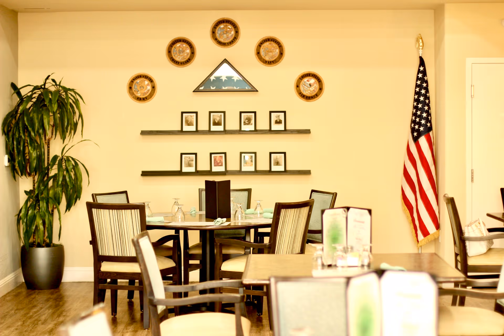 Dining room with round tables and chairs, wall-mounted framed photos and plaques, a potted plant, and an American flag.