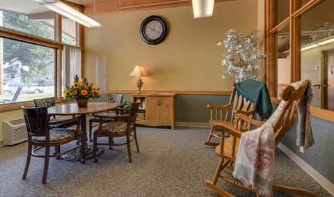 A cozy interior room with a round wooden table surrounded by four chairs with floral cushions. To the right, there are two wooden rocking chairs draped with blankets. A wooden sideboard with a lamp and a clock on the wall above it is visible. Large windows on the left let in natural light and show a view of trees and parked cars outside.