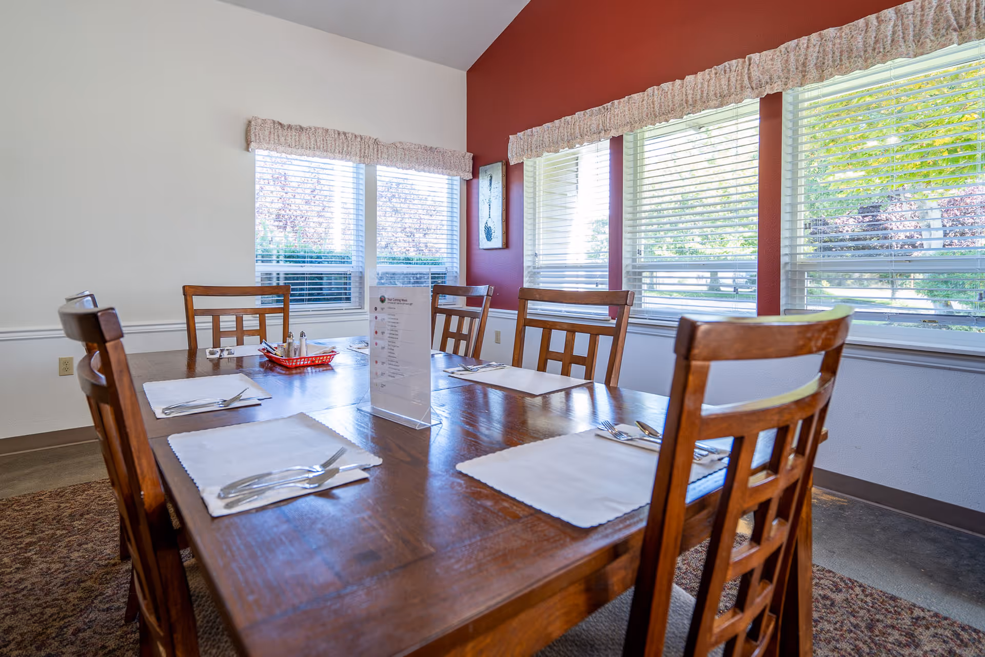 Sunlit dining room with a wooden table set with placemats and utensils and chairs by large windows.