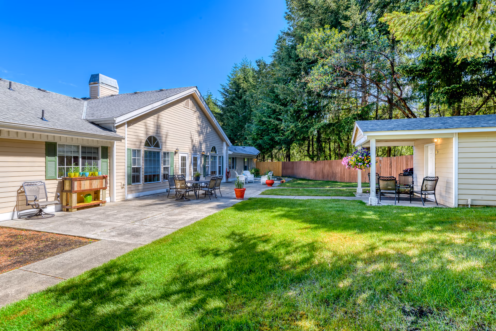 Outdoor view of Hearthside Manor Memory Care facility showing a beige building with large windows and green shutters, a concrete patio with outdoor chairs and tables, a small covered seating area with chairs, green grass lawn, potted plants, and trees in the background under a clear blue sky.