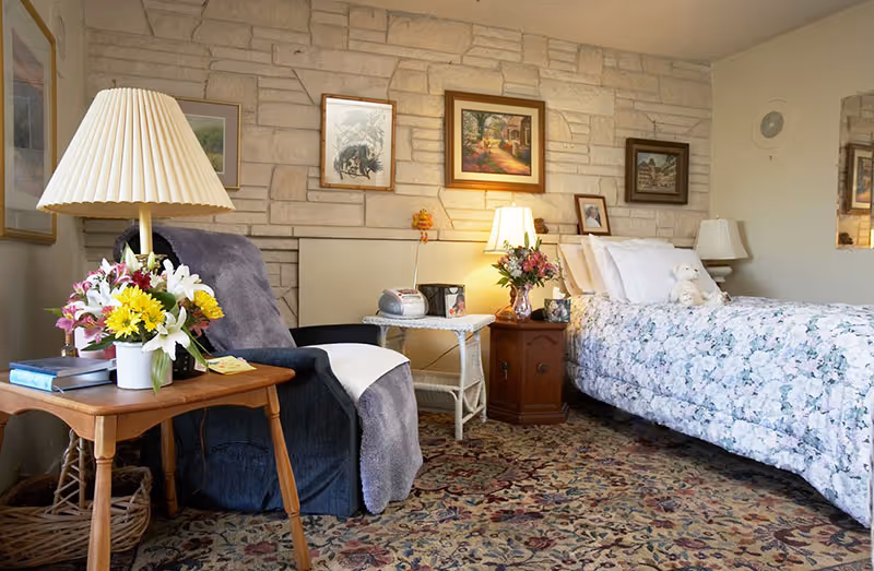 A cozy bedroom in an assisted living facility featuring a single bed with a floral bedspread and a teddy bear on the pillow. Next to the bed is a wooden nightstand with a lamp and a vase of flowers. A comfortable armchair with a gray throw blanket is positioned beside a wooden side table holding a lamp, a bouquet of flowers, and some books. The room has a patterned carpet and a stone accent wall adorned with framed artwork.