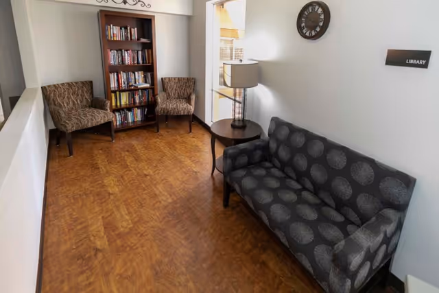 A small library room with wooden flooring, featuring a dark patterned sofa on the right, two patterned armchairs on the left, a wooden bookshelf filled with books against the back wall, a round wooden side table with a lamp, a wall clock, and a sign labeled 'Library'.