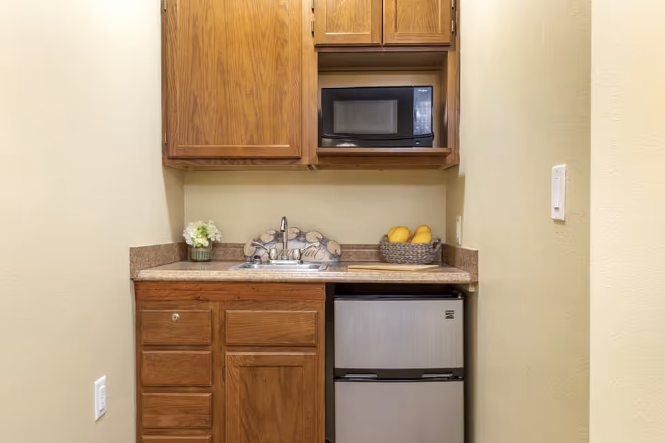 Small kitchenette with wooden cabinets, a microwave, a sink, a mini refrigerator, a basket with yellow fruits, and a small flower vase on the countertop.