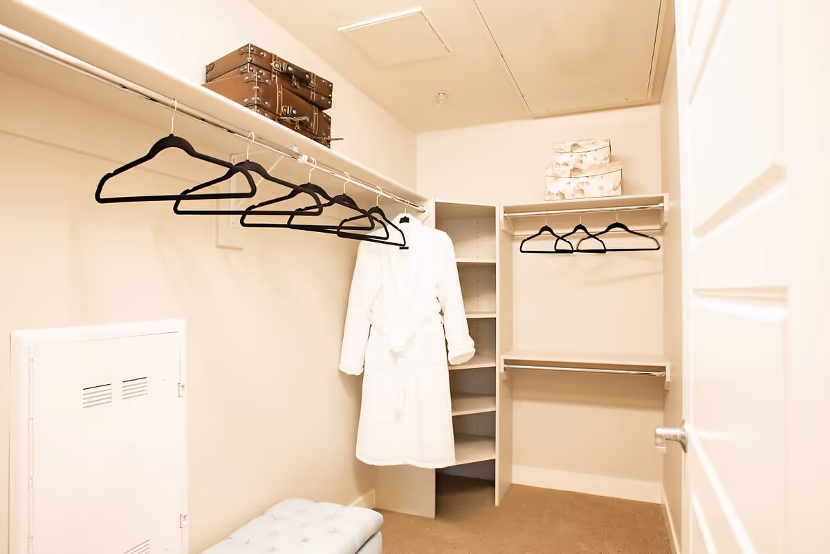 A walk-in closet with beige walls and carpeted floor. There are two shelves with hanging rods and several black hangers. A white bathrobe is hanging on one of the rods. On the top shelves, there are two decorative suitcases and three floral-patterned storage boxes. A small cushioned bench is visible at the bottom left corner.