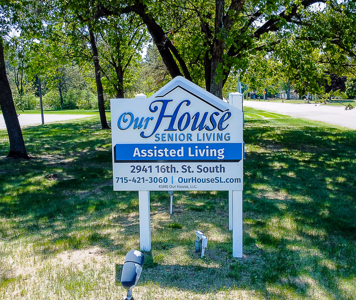 Outdoor view of a white sign for Our House Senior Living Assisted Living located on a grassy area with trees and a road in the background.