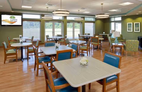 A bright and spacious dining room with multiple tables and chairs arranged neatly. The room has large windows allowing natural light to fill the space, green walls, and modern ceiling lights. A television mounted on the wall displays the Lorien Bulle Rock logo. There are small flower arrangements on the tables and comfortable seating areas with armchairs in the background.