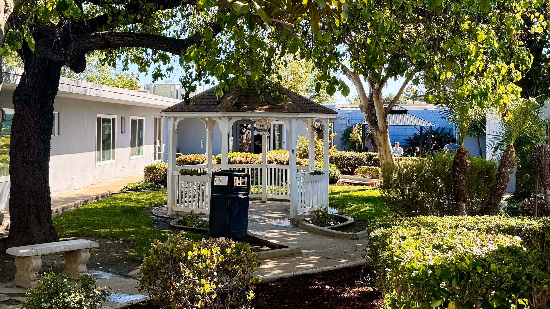 A peaceful outdoor garden area at Brighton Place Spring Valley featuring a white gazebo surrounded by green grass, bushes, and trees. There is a stone bench on the left side and a trash can near the gazebo. In the background, part of the building and a few people sitting can be seen.