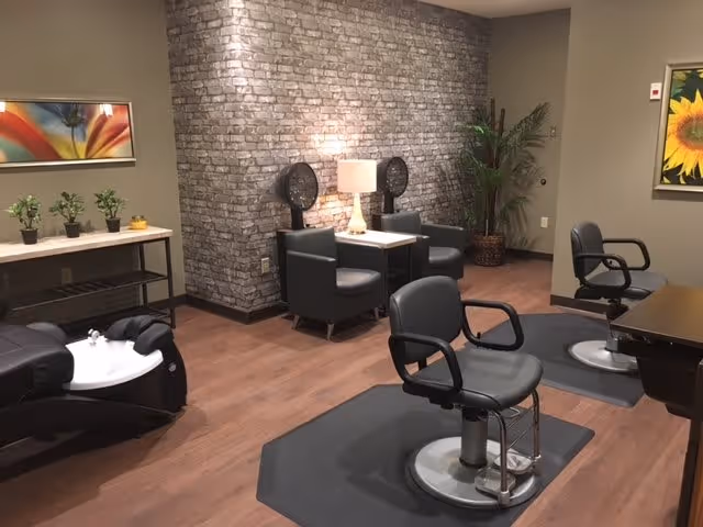 Interior of a salon area in a senior living facility with two salon chairs on mats, a hair washing station, two black chairs with hair dryers, a table with a lamp, potted plants, and colorful wall art.