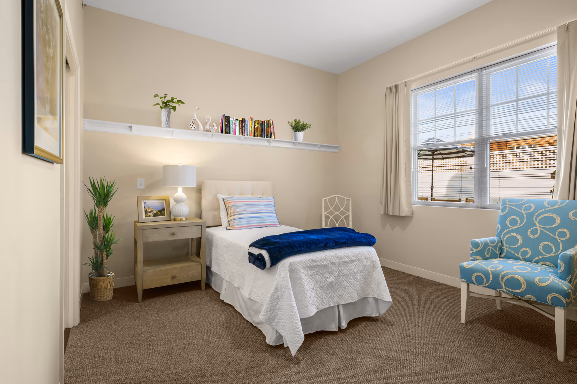 A neatly arranged bedroom with a single bed covered in white bedding and a blue blanket folded at the foot. Next to the bed is a wooden nightstand with a white lamp, a framed photo, and a small plant. Above the bed is a white shelf holding books and decorative items. A blue armchair with a circular pattern is placed near a window with beige curtains, letting in natural light. The room has beige walls and carpeted floor.
