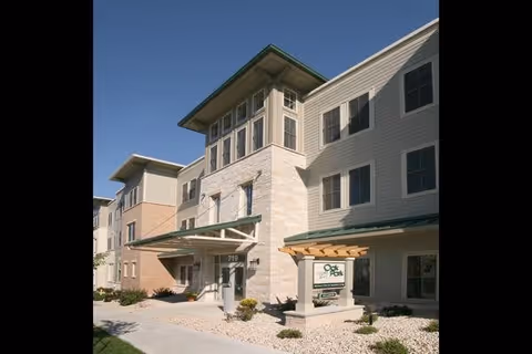 Exterior view of a multi-story senior living facility building with beige and light brown siding, large windows, and a covered entrance. There is a sign near the entrance that reads 'Oak Park Place Madison'. The landscaping includes small bushes and rocks along the walkway leading to the entrance.