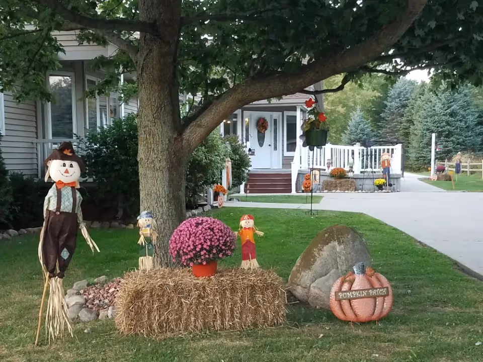 House front yard decorated for fall with scarecrows, a hay bale topped with pink mums, a pumpkin labeled "Pumpkin Patch", and a tree in front of the porch.