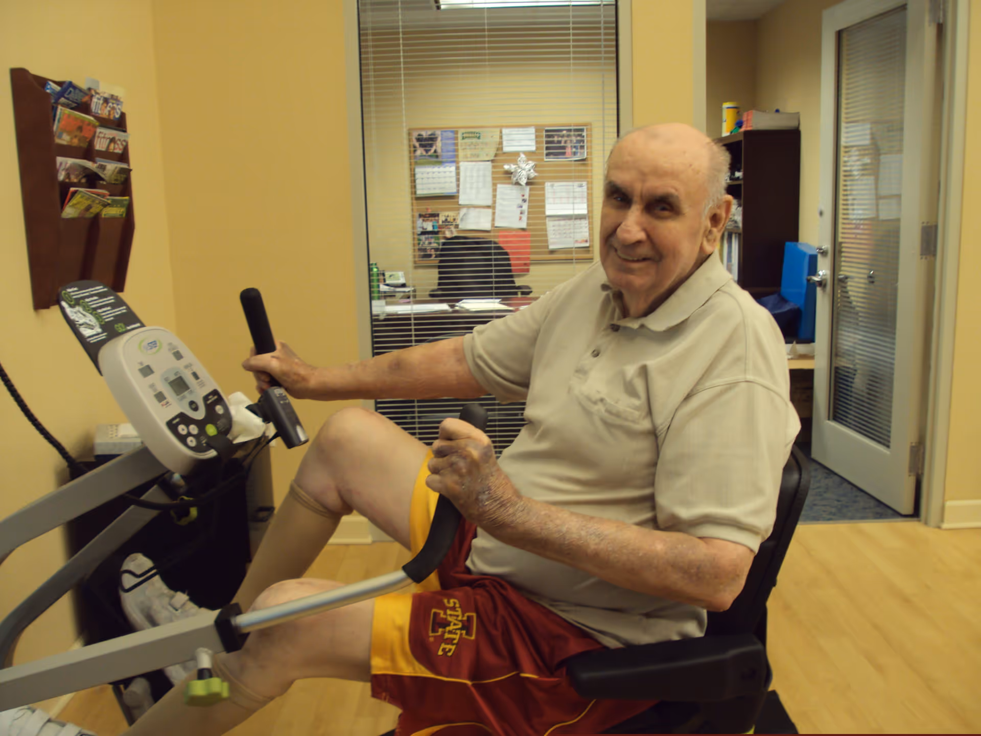 An elderly man wearing a beige polo shirt and red and yellow shorts is seated on a stationary exercise bike in a room with yellow walls and wooden flooring. He is smiling and holding the bike's handles. Behind him, there is a glass window with blinds showing an office area with a bulletin board and desk. A magazine rack is mounted on the wall to the left.