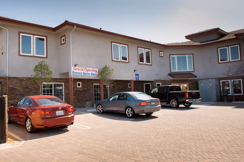 Exterior view of Ramona Senior Manor showing a two-story building with multiple windows and a stone facade on the lower part. There are three vehicles parked in front on a paved parking area. A banner on the building reads 'Grand Opening Ramona Senior Manor'.