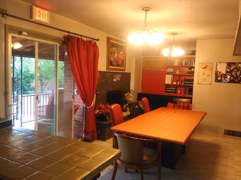 A warmly lit dining room with a long rectangular table and chairs, chandeliers overhead, red curtains at a sliding glass door, and a fireplace with shelving in the background.