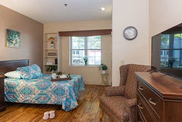 A cozy bedroom in a senior living facility with a single bed covered in a blue and green patterned bedspread. There is a wooden nightstand with a lamp next to the bed, a pair of pink slippers on the wooden floor, a cushioned armchair with a patterned fabric, a wooden dresser with a flat-screen TV on top, and a window with a view outside. The walls are painted beige and light brown, and there is a clock on the wall above the armchair.