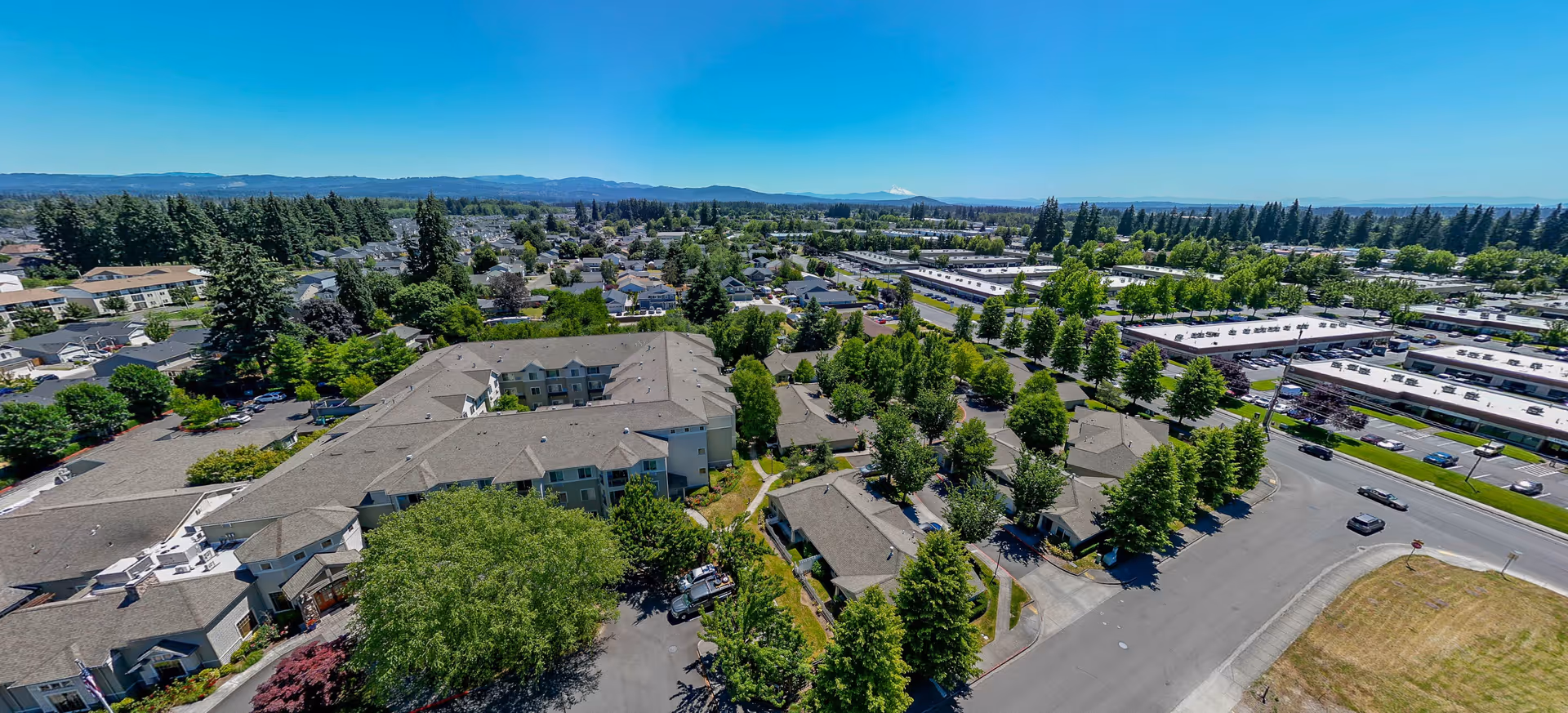 Aerial view of Brookdale Vancouver Orchards senior living facility and surrounding neighborhood with multiple buildings, trees, roads, and parked cars under a clear blue sky.